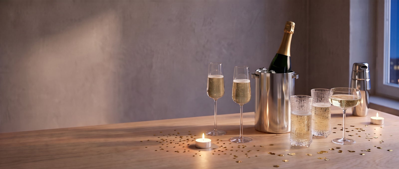 Iittala Essence flutes and Ultima Thule glasses with sparkling wine, a Stelton AJ champagne cooler with a bottle, a Blomus Belo coupe and an Alessi 870 shaker on an oak table with gold confetti